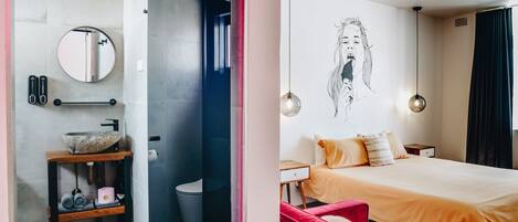 A modern bathroom with a round mirror, a vessel sink, and a towel rack. The room features a large bed with a red chair, a wall-mounted light fixture, and a painting.