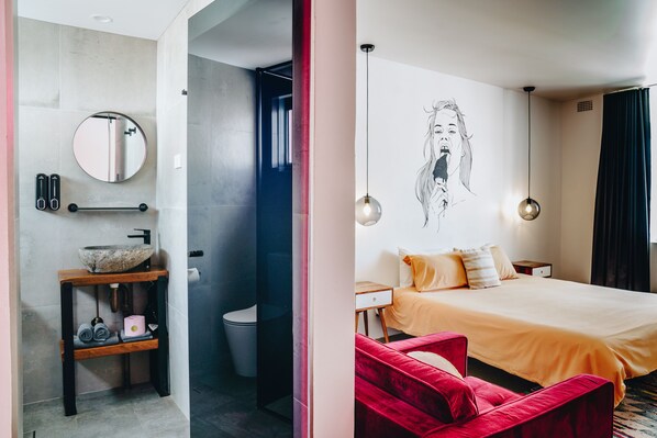 A modern bathroom with a round mirror, a vessel sink, and a towel rack. The room features a large bed with a red chair, a wall-mounted light fixture, and a painting. - The Pink Hotel Coolangatta (Coolangatta)