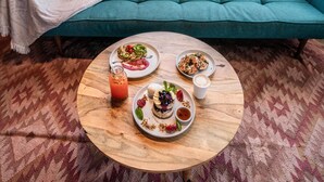 Een smaragdgroene bank met een witte deken en een enkel smaragdgroen kussen, een ronde houten salontafel met schotels eten en drinken, en uitzicht op een park met bomen en een heldere lucht door een raam.