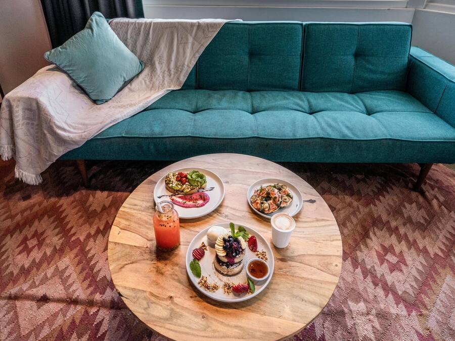 Een smaragdgroene bank met een witte deken en een enkel smaragdgroen kussen, een ronde houten salontafel met schotels eten en drinken, en uitzicht op een park met bomen en een heldere lucht door een raam.