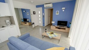 Una sala de estar moderna con un sofá azul, una mesa de centro y un televisor. Se observa una cocina con un fregadero y armarios.