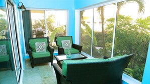 Una habitación con un gran ventanal corredero que da a un balcón, mobiliario de mimbre con cojines verdes y vistas a un follaje exuberante.