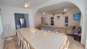 Un comedor amplio con una mesa larga y sillas blancas, un ventilador de techo y un televisor montado en la pared.