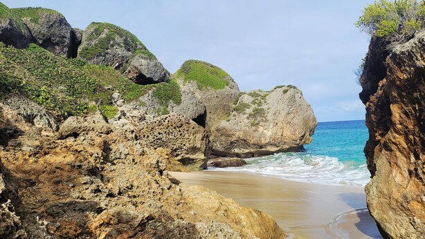Un paisaje costero rocoso con un estrecho paso entre grandes rocas que conduce a una playa de arena.