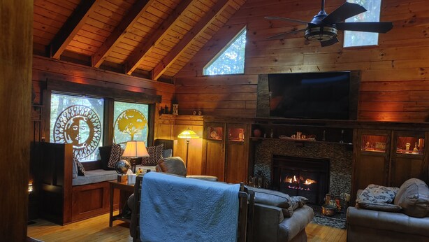 Interior acollidor d'una cabana amb una llar de foc, parets de fusta, un ventilador de sostre i una finestra gran.