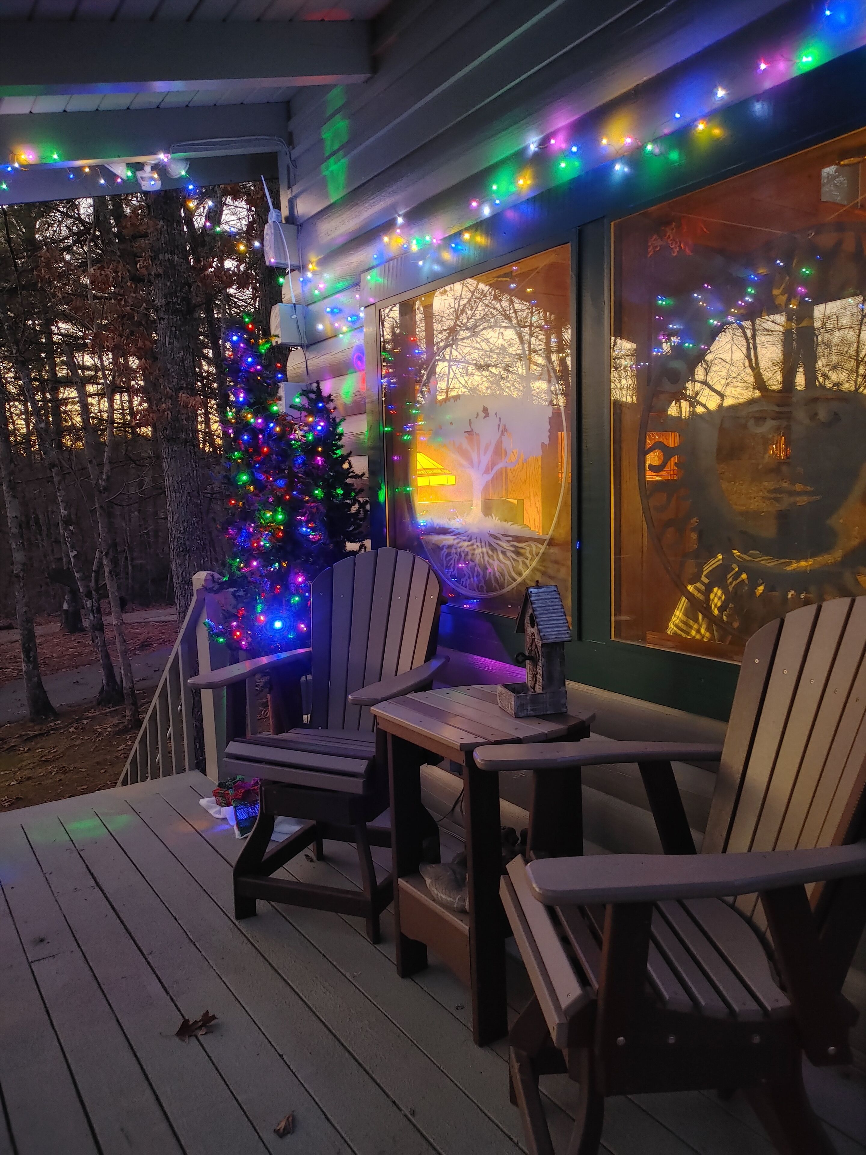 A wooden deck with two Adirondack chairs and a small table, decorated with colorful string lights and a Christmas tree.
