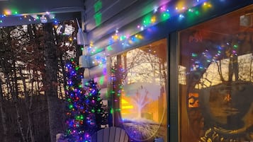 A wooden deck with two Adirondack chairs and a small table, decorated with colorful string lights and a Christmas tree.