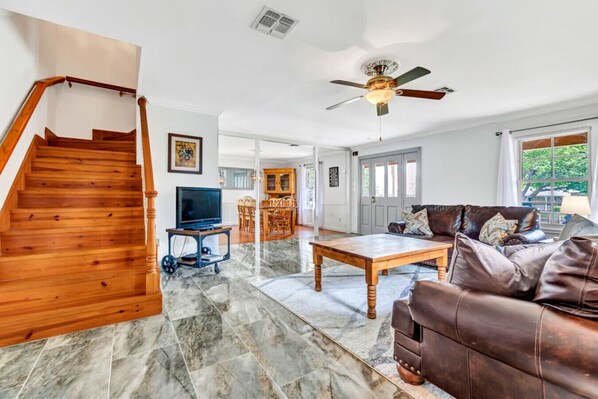 A spacious living area with a wooden staircase, a ceiling fan, a television, a dining area, and a leather sofa. - Ladies Castle on Main; 14 Beds (Fredericksburg)
