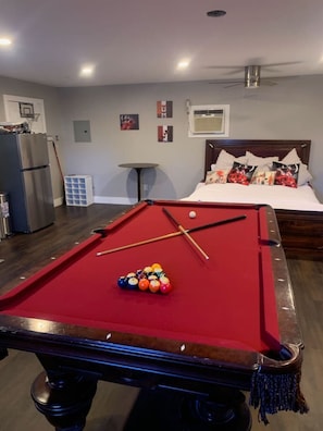 A pool table with balls, a bed with pillows, a ceiling fan, and a refrigerator.