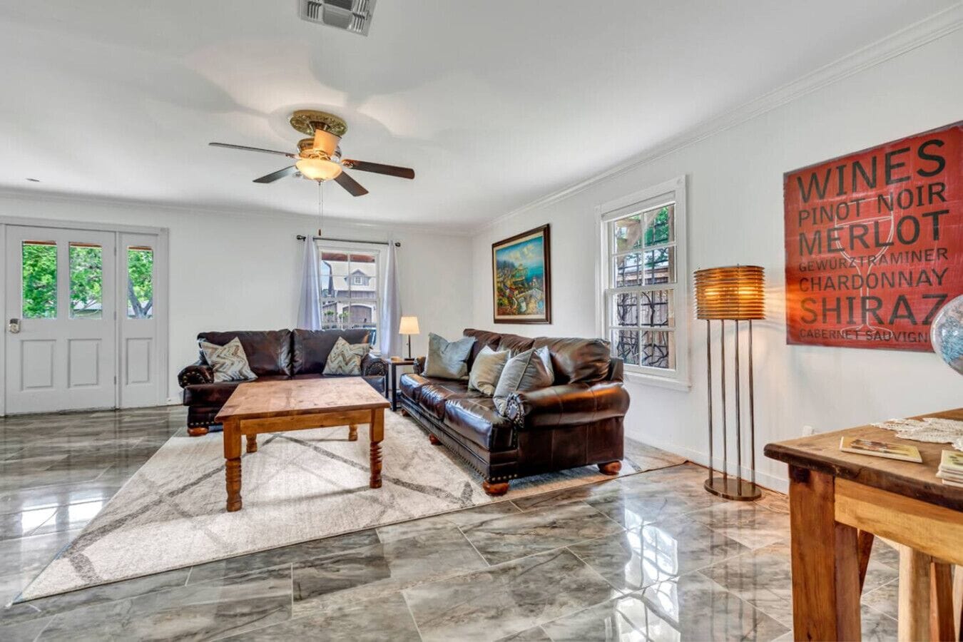 A spacious living room with a dark leather sofa, a wooden coffee table, and a floor lamp. There is a painting on the wall and a large window with curtains.