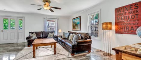 A spacious living room with a dark leather sofa, a wooden coffee table, and a floor lamp. There is a painting on the wall and a large window with curtains.