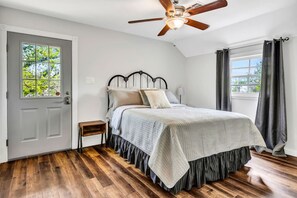 Quarto com piso de madeira, cama com cabeceira, mesa de cabeceira, ventilador de teto e janela com cortinas.