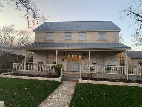 A two-story stone house with a front porch, a stone pathway, and a fenced yard. - Ladies Castle on Main; 14 Beds (Fredericksburg)
