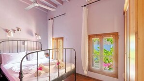 Een slaapkamer met een metalen bedframe, een houten vloer, een raam met uitzicht op groen en een plafondventilator.