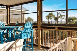 Overdekt terras met blauwe Adirondack stoelen en een houten tafel, met uitzicht op een strand met palmbomen en een heldere lucht.