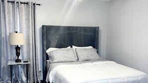 Ein Schlafzimmer mit einem Bett, einem Deckenventilator, einem Nachttisch mit Lampe und einem mit Vorhängen versehenen Fenster.