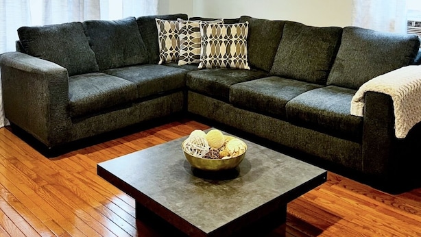 Ein Wohnzimmer mit einem dunkelgrünen Ecksofa, gemusterten Zierkissen, einem Couchtisch mit einer Schale dekorativer Gegenstände, Holzböden und weißen Vorhängen.