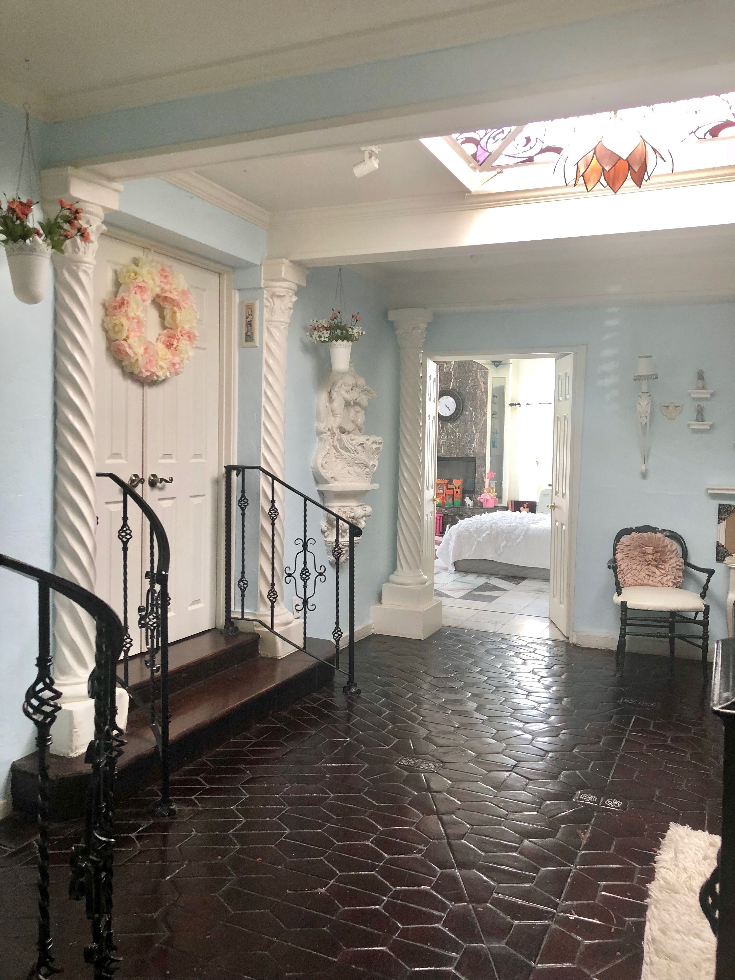 A foyer with a stone floor, a staircase with ornate black railings, a white door with a floral wreath, and a view into a bedroom with a bed and a dresser.