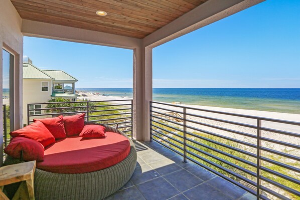 A balcony with a wicker sofa and red cushions, overlooking a beach and ocean.