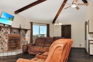 A living room with a stone fireplace, a large flat-screen TV, a ceiling fan, and a view of the outdoors.