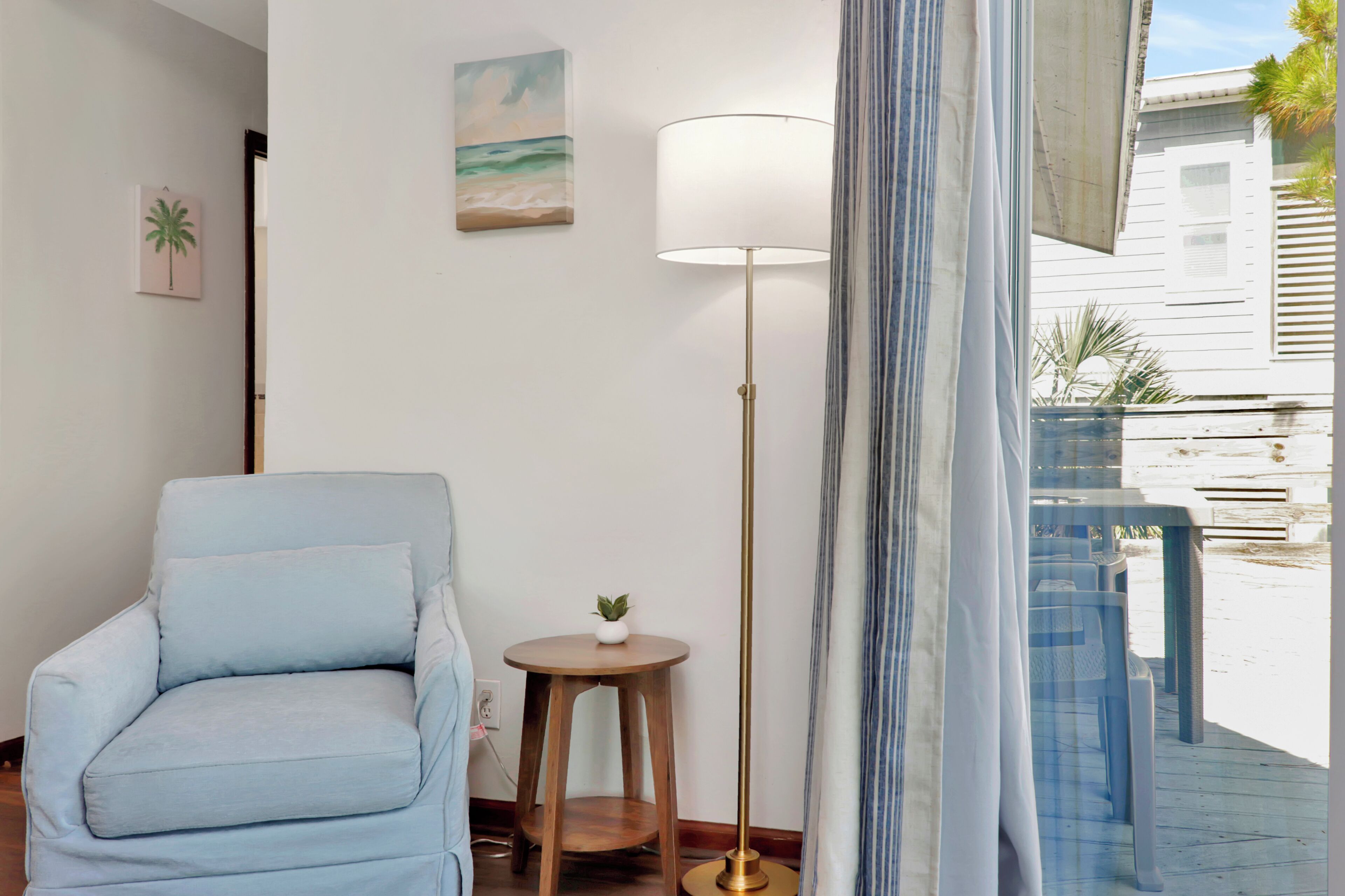A room with a light blue armchair, a small round wooden table with a plant, a tall floor lamp, and a beach-themed painting on the wall.