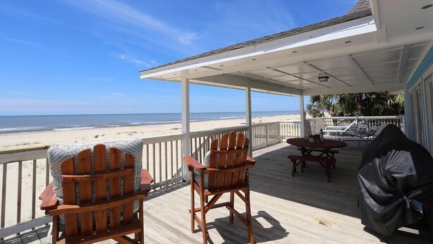 Una terraza de madera con dos sillas Adirondack, una mesa de picnic y una parrilla, con vista a la playa.