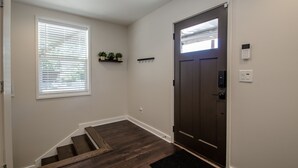 A hallway with a dark wooden floor, a white door with a peephole, a window with blinds, and a small shelf with plants. - NEW Modern House with Roof Deck of Atlanta Skyline (Atlanta)