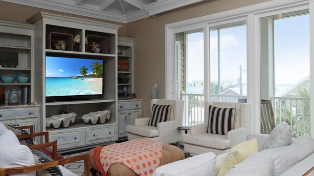 A living room with a television displaying a beach scene, white furniture, and a balcony with a view. - Gulf View, Large Home with Elevator, and Easy Beach Access ~ Sammys Old Place (Indian Pass)