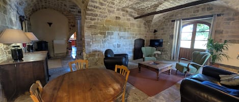 A stone-walled room with a round wooden table and chairs, a sofa, and a television.
