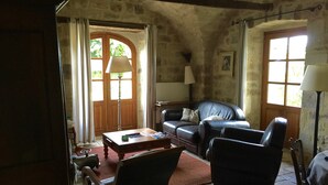 A rustic living room with a leather sofa, a wooden coffee table, and a stone floor. There is a door leading outside, and a lamp is visible on the right side. - Renovated farmhouse of the 16th, charming, south Ardèche (Chassiers)
