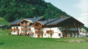Un bâtiment de style chalet, doté de nombreux balcons et d’un toit en pente, entouré d’une zone herbeuse et d’une colline boisée.