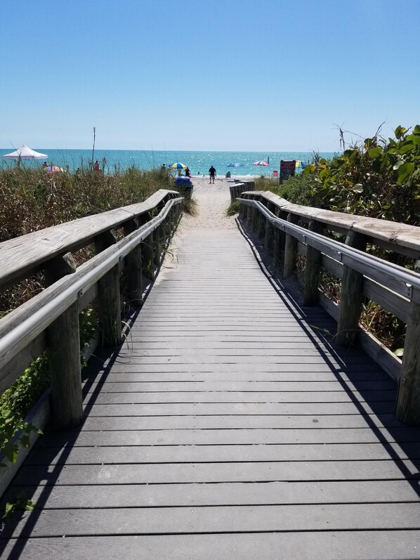 A wooden boardwalk leading to a beach with umbrellas and people in the distance.