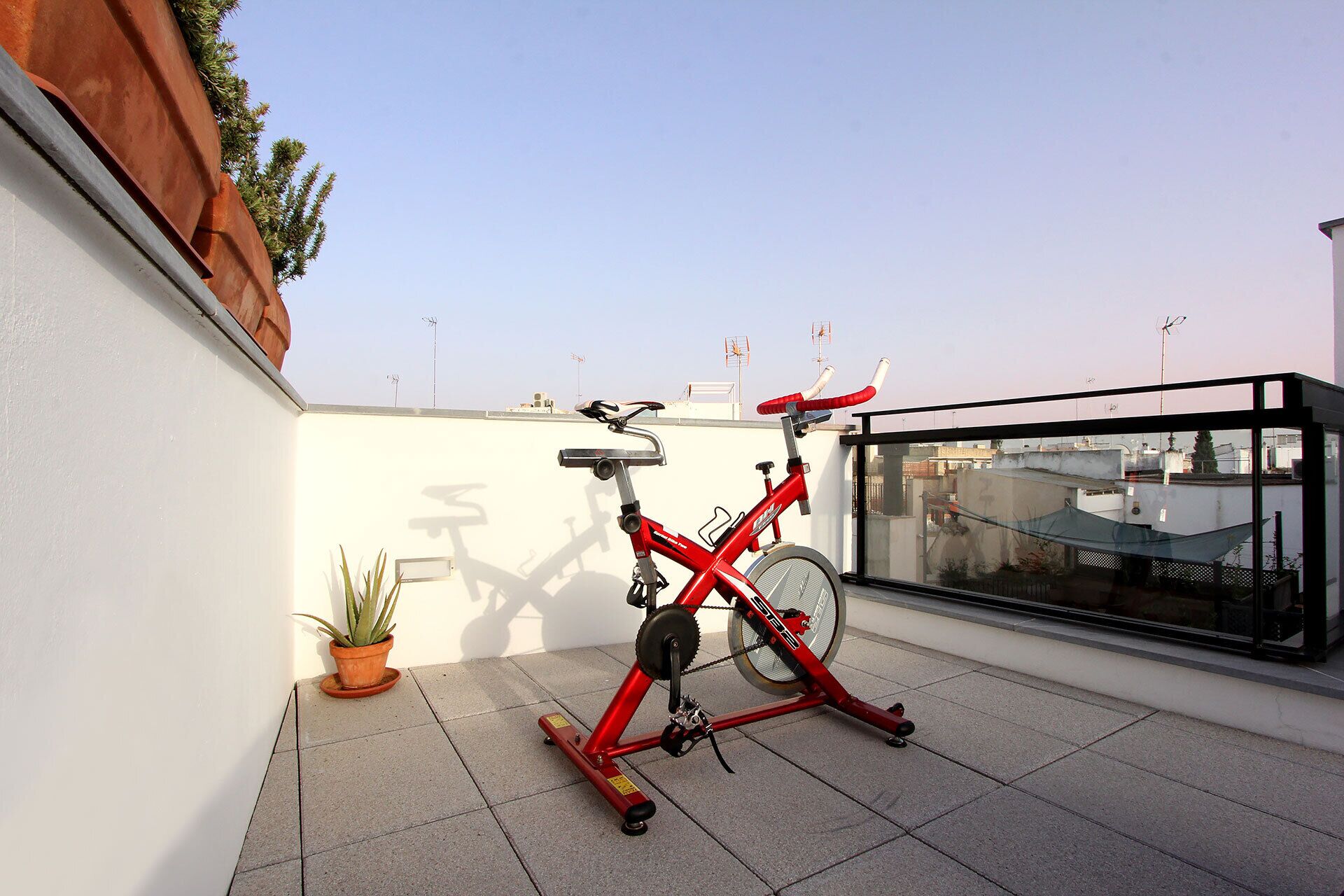 Una cyclette rossa su un balcone piastrellato con una pianta in vaso e una struttura in vetro.