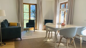 Uma sala de estar moderna com um sofá, uma mesa de jantar e cadeiras. Há uma televisão em um suporte, um abajur e uma janela com cortinas.