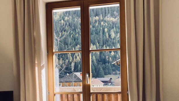 Quarto com piso de madeira, banheira branca e uma grande janela com vista para uma área arborizada.