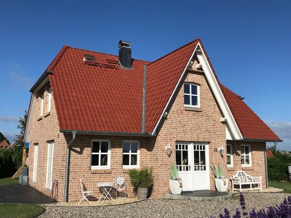 Une maison à deux étages, construite en briques, avec un toit recouvert de tuiles rouges, un porche à l’avant doté d’un banc et un petit jardin.