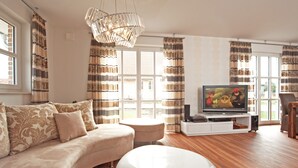 Un salon moderne avec un canapé d’angle, une table basse blanche, un téléviseur posé sur un meuble blanc et une grande fenêtre avec des rideaux.