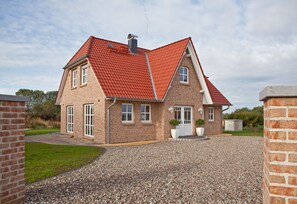 Une maison à deux étages, avec un toit recouvert de tuiles rouges, une allée en gravier et une façade en briques.
