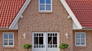 Une maison en briques avec un toit recouvert de tuiles rouges, une porte blanche et deux plantes en pot sur le porche.