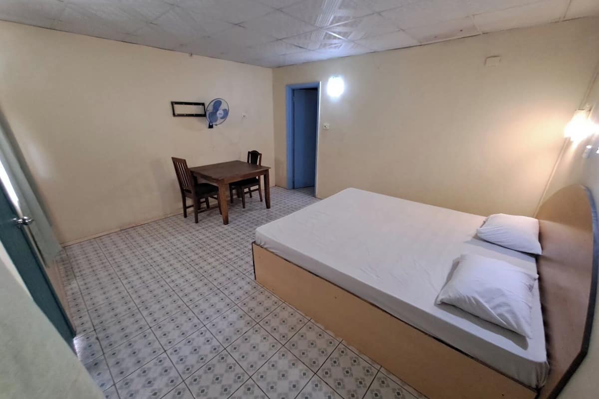 Una habitación pequeña y limpia con una cama individual, una mesa con sillas y un ventilador en la pared.