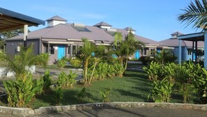 A hotel with multiple buildings, each with a grey roof and blue doors. There are lush green plants and a well-maintained lawn in front. - Fun World Plaza Hotel (Nadi)