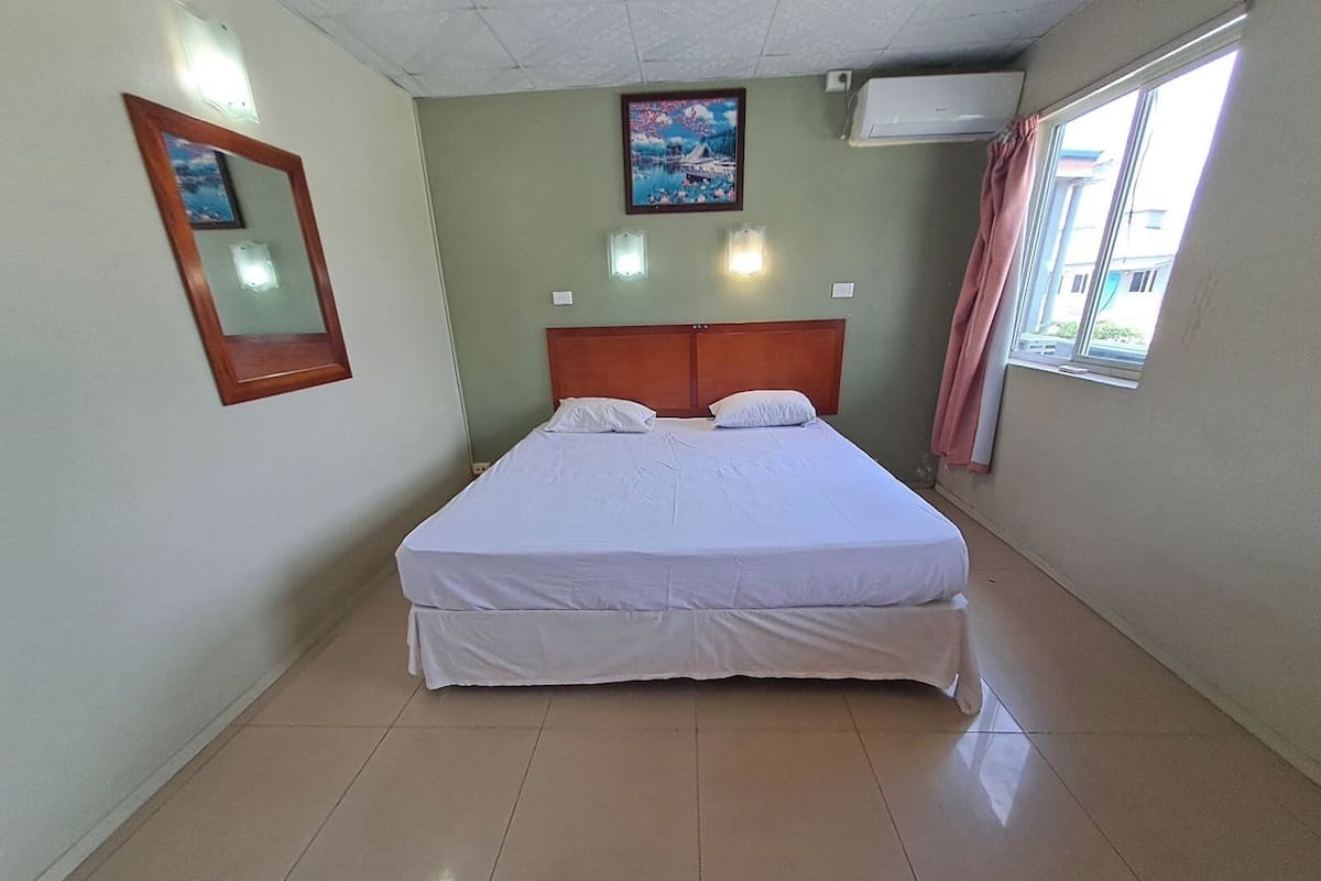 Habitación de hotel limpia y bien iluminada con una cama grande, cabecera, aire acondicionado instalado en la pared, ventana con cortinas y un cuadro sobre la cama.