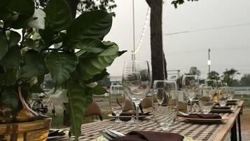 An outdoor dining table set with plates, glasses, and cutlery, surrounded by trees and string lights.