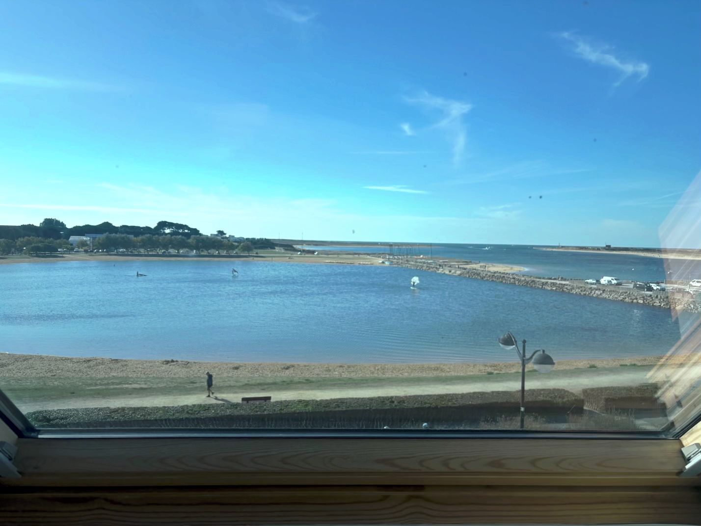 Vue depuis l’intérieur d’un bâtiment, donnant sur une étendue d’eau avec une plage de sable et une digue rocheuse.