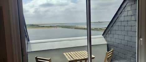 Ein Balkon mit Blick auf ruhiges Wasser, einen Sandstrand und eine entfernte Küstenlinie. Auf dem Balkon stehen Holzstühle und ein Tisch.