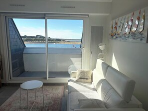 Un balcon avec vue sur une plage et la mer, un canapé blanc, une petite table ronde et un tapis à motifs.