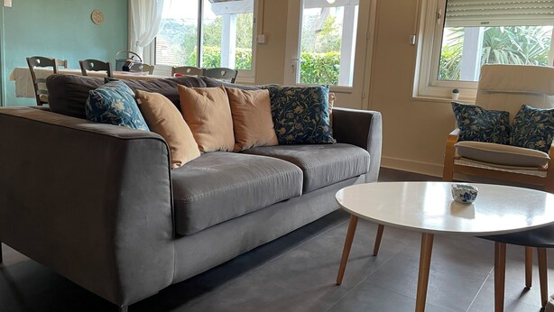 A living room with a grey sofa, a round coffee table, and a dining area with chairs and a table. - 3* Gîte l'ancienne Epicerie Meyronne (Meyronne)