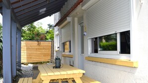 A covered patio area with a wooden bench and a table, adjacent to a building with white siding and a window with blinds. - 3* Gîte l'ancienne Epicerie Meyronne (Meyronne)