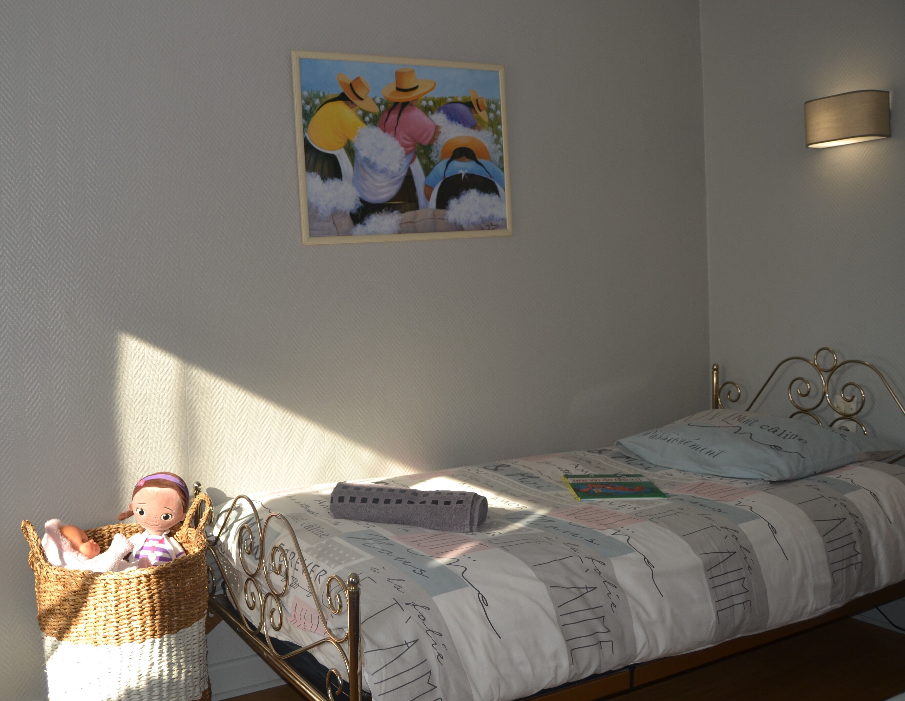 Une chambre à coucher avec un lit, une lampe fixée au mur, un tableau et un panier contenant une poupée.