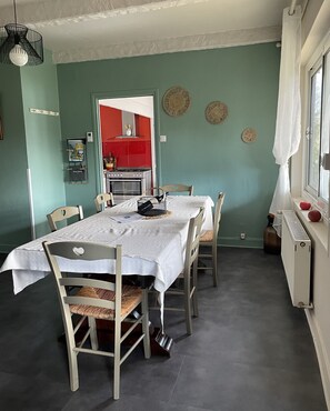 Un comedor con una mesa dispuesta para cuatro personas, rodeada de sillas de madera con asientos de mimbre. Se ve una cocina roja a través de una puerta abierta al fondo.